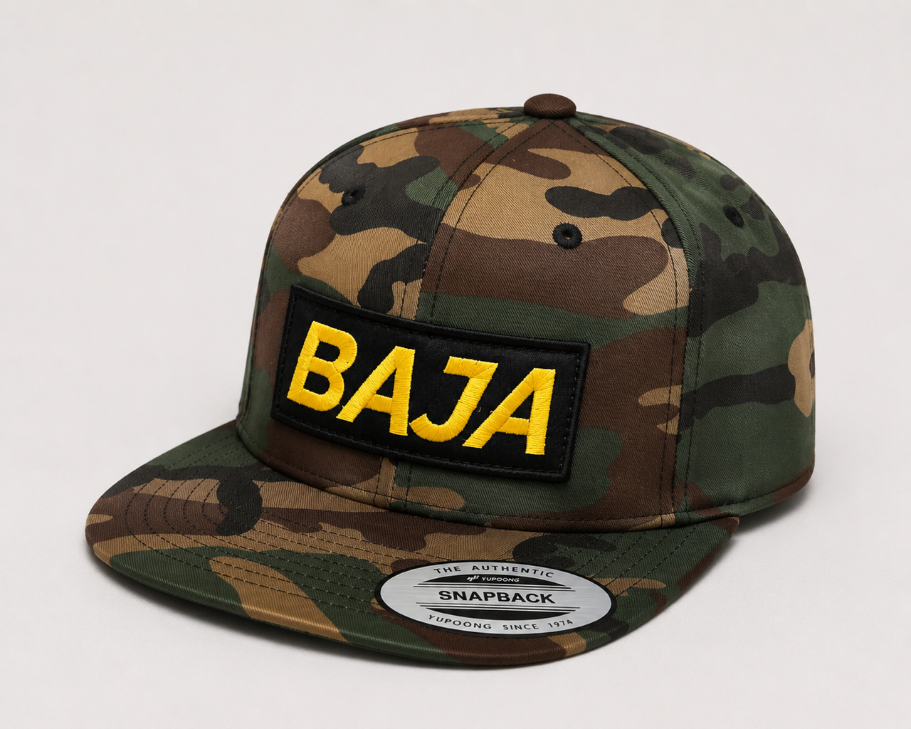A camouflage snapback cap with a black rectangular patch on the front displaying 'BAJA' in bold yellow letters. The cap features a classic woodland camo pattern in green, brown, and black, with a visible authentic snapback sticker on the flat brim.