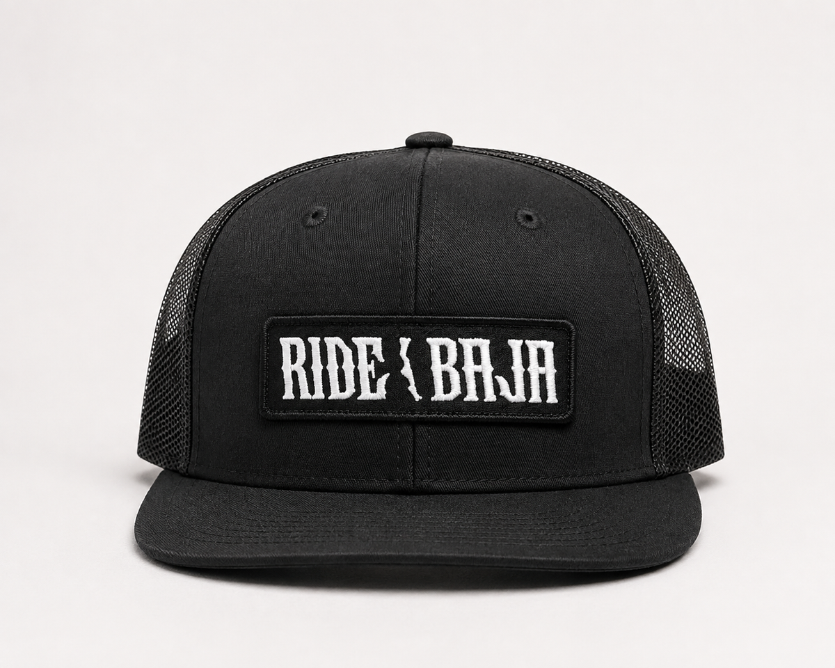 A black trucker-style cap with a black mesh back and a rectangular front patch reading 'RIDE OR BAJA' in white gothic lettering, photographed on a clean white background.