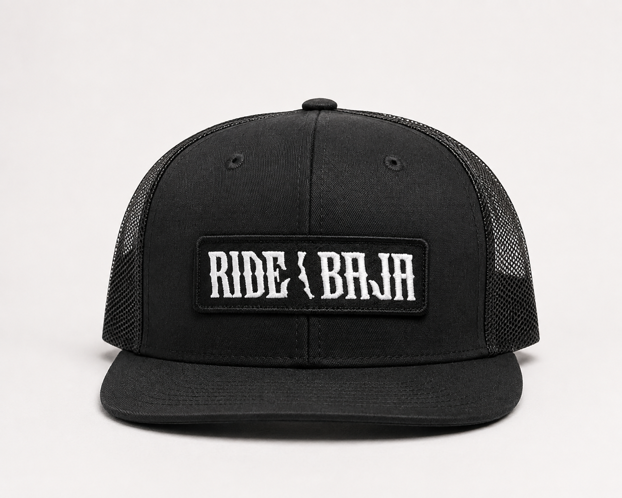 A black trucker-style cap with a black mesh back and a rectangular front patch reading 'RIDE OR BAJA' in white gothic lettering, photographed on a clean white background.